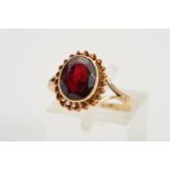 A GARNET RING, designed as an oval shape garnet in a collet setting within bifurcated shoulders on a