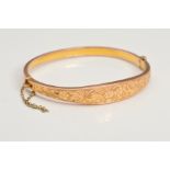 A 1920'S 9CT GOLD HINGED BANGLE, featuring a floral engraved front panel, with a safety chain,