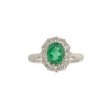 An emerald and diamond cluster ring, the oval emerald within a brilliant-cut diamond surround,