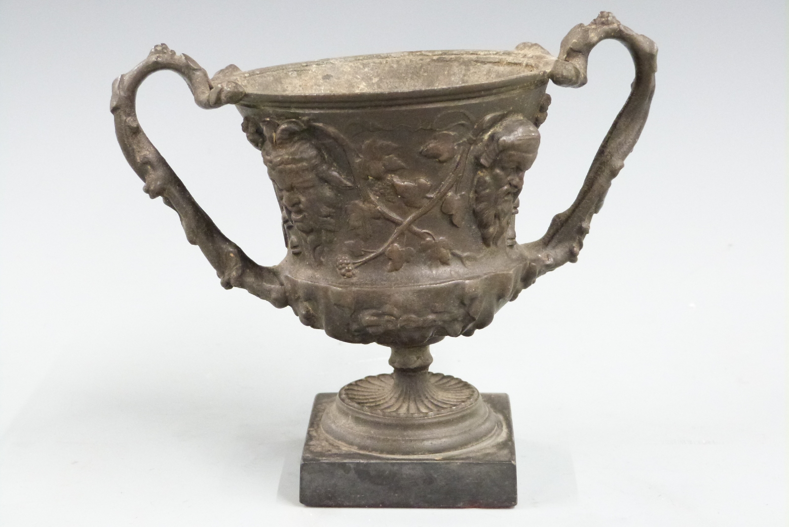 Spelter miniature twin handled vase or urn with mask and foliate decoration, on square slate or