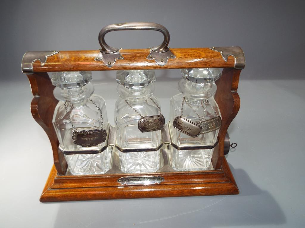 An oak and white metal Tantalus, holdfast by Walker & Hall, fitted with three cut glass decanters,