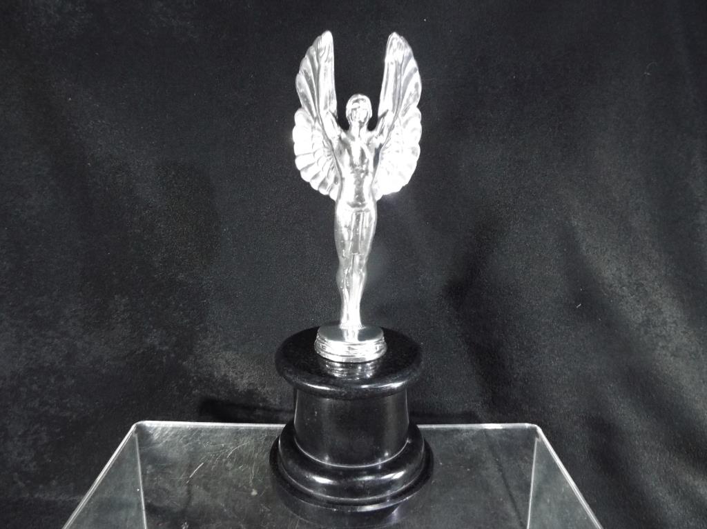 A white metal figure depicting a winged angel mounted on a circular black plinth,