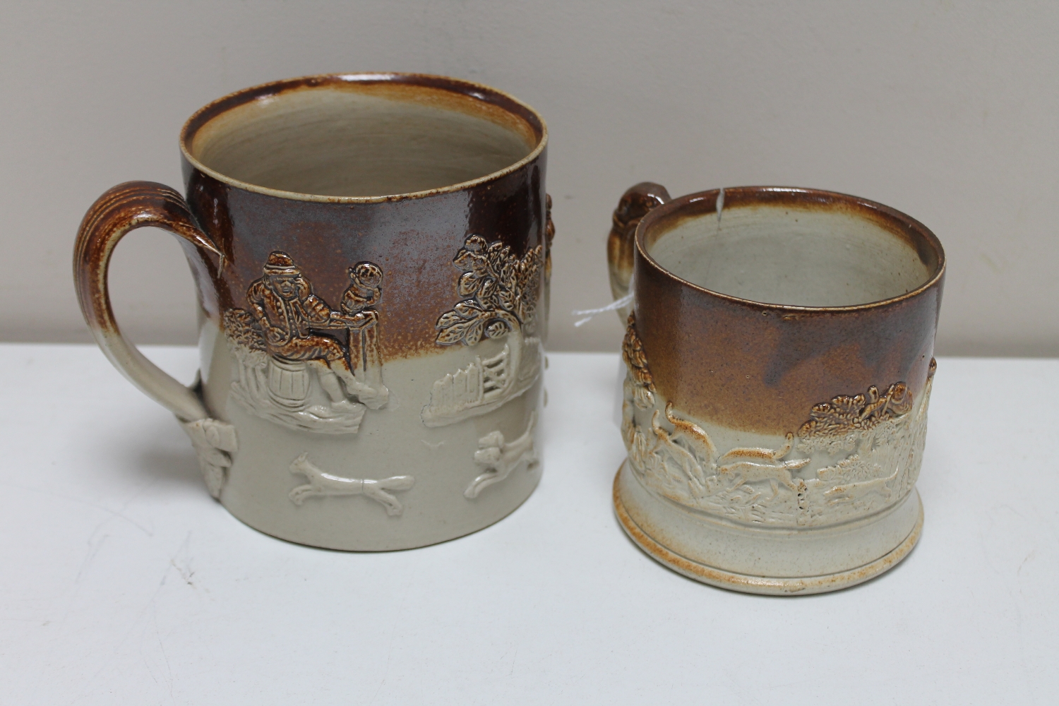 Two antique stoneware mugs depicting hunting and farming scenes.