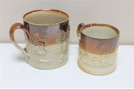 Two antique stoneware mugs depicting hunting and farming scenes.