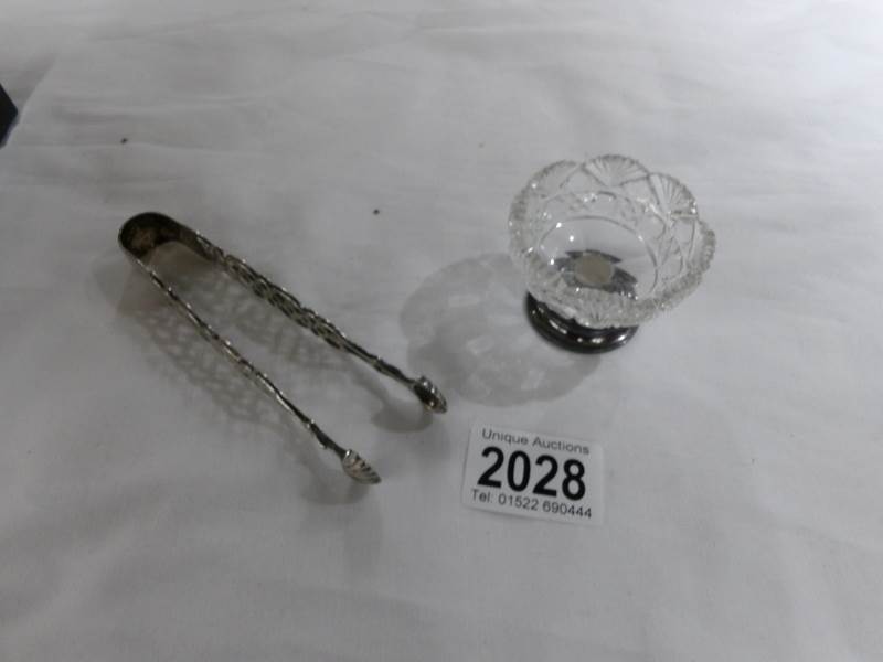 A pair of silver sugar tongs and a glass salt with silver base.