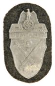 A Third Reich Demjansk shield, of zinc plated iron on field grey cloth patch with original paper