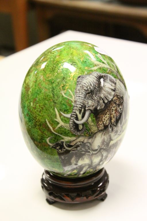 Hand painted signed ostrich egg on carved hardwood stand, green and gilt ground depicting lioness
