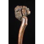 A Rustic Root-wood Stick having a burr to one end carved with a face, 25" (63.5 cm) in length.