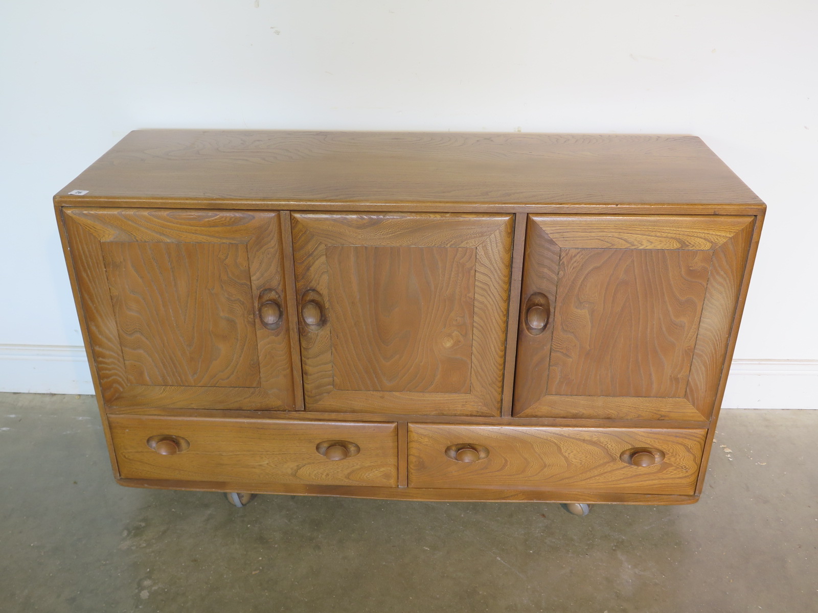 An Ercol blonde elm and beech sideboard with three cupboard doors over three drawers, missing its