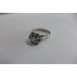 A white metal cluster ring set with a diamond and six emeralds, centre brilliant cut diamond is held