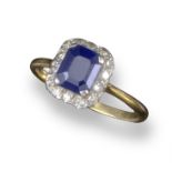 A sapphire and diamond cluster ring, the emerald-cut sapphire is set within a surround of single-cut
