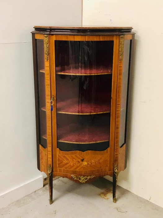 An Ornate corner cabinet with brass detailing, marquetry and a curved glass door and three shelves.