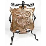 An Arts and Crafts iron framed fire screen with copper central panel relief decorated with a dragon,