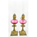 A small pair of brass oil lamps with cranberry glass reservoirs and clear glass chimneys, height