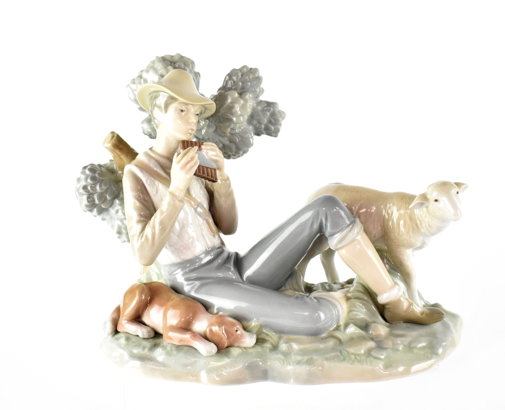 A Lladró figure of a young shepherd boy seated on a grassy knoll playing pan pipes,