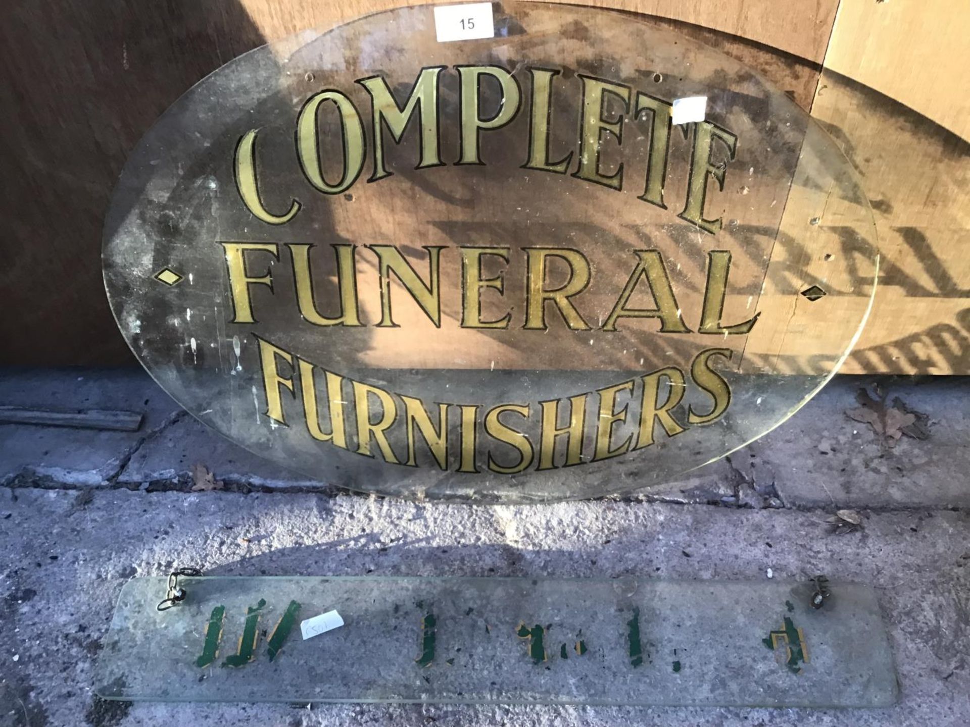 AN OVAL GLASS SIGN PAINTED WITH 'COMPLETE FUNERAL FURNISHERS' AND A FURTHER GLASS HANGING PLAQUE (