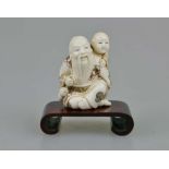 Netsuke, Japan, wohl 19. Jh., sitzender, alter Mann mit Kind auf der Schulter, Elfenbein, am Boden