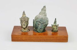 Drei abgeschlagene Köpfe ehemaliger Tempelfiguren, Südostasien, Bronze, auf Holzsockel mit Kn