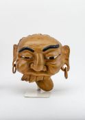 HolzmaskeHolz, geschnitzt. Grimasse schneidendes Gesicht, mit Ohrringen, geöffnetem Mund mit vier