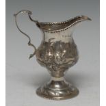 A George III silver pedestal cream jug, embossed and engraved with foliate scrolls,