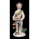 A Bow figure of a putto: holding a basket of flowers and wearing a floral headdress and sash,