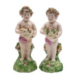 Two Derby figures of putti: each holding a basket of flowers, on green mound bases, patch marks,