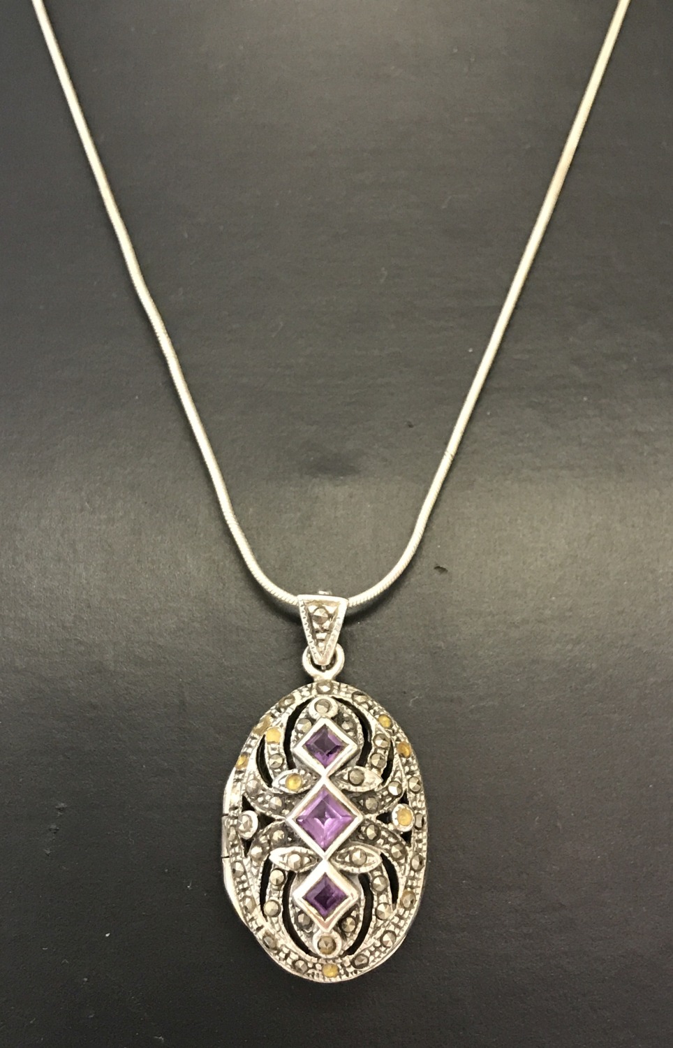 A silver oval locket with pierced work detail to front. Set with square cut amethysts & marcasite's.