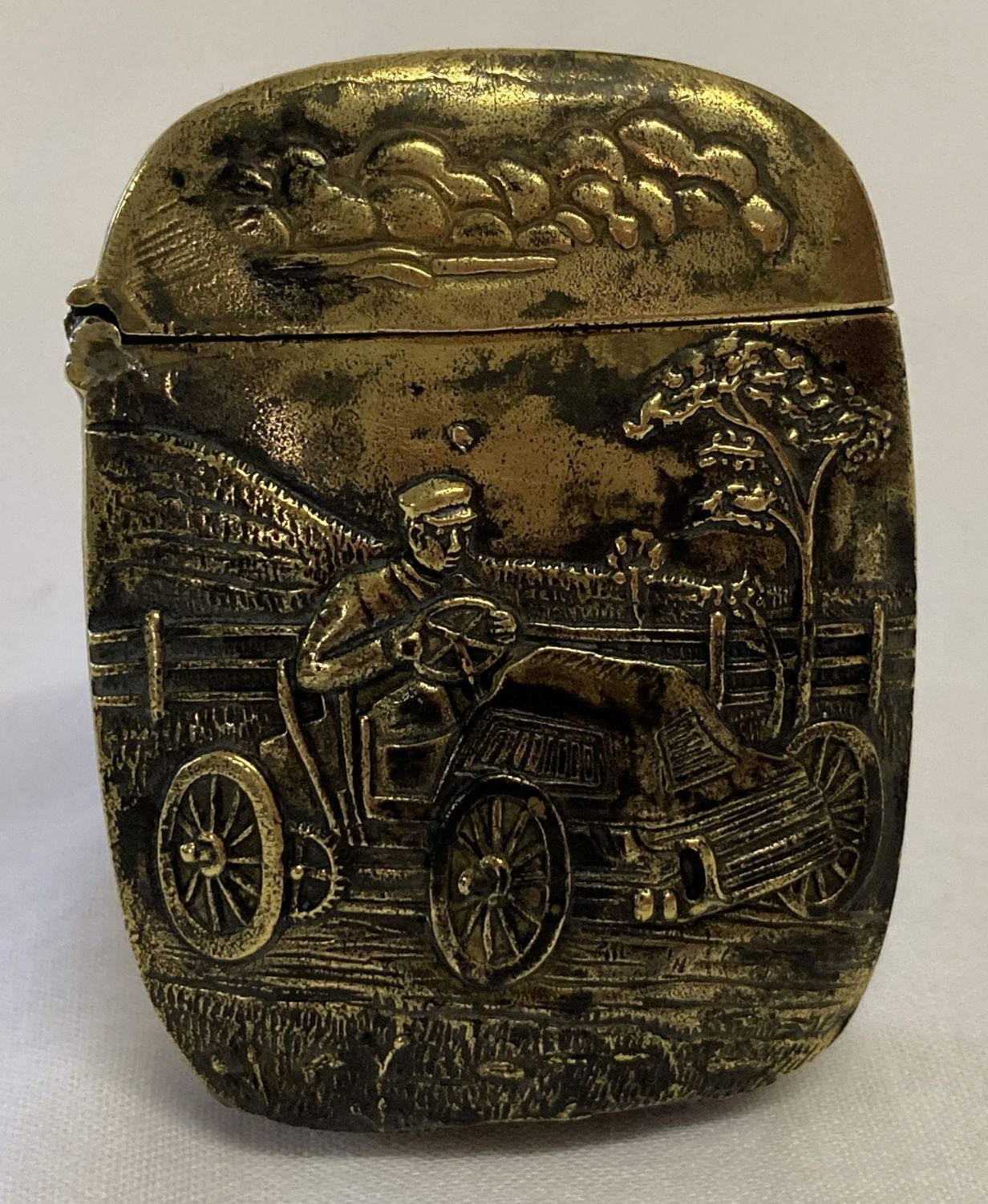 A brass vesta case featuring a man driving a car. Detailing to both sides.