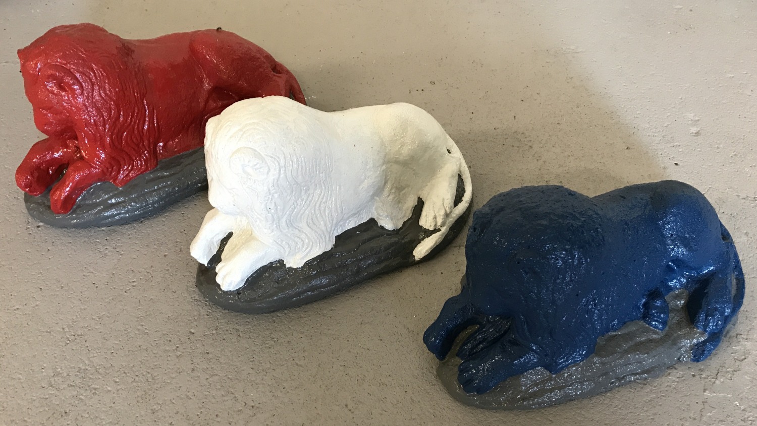 3 stone garden ornaments in the shape of laying lions, painted in red, white and blue colours.