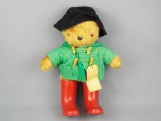 A Paddington Bear soft toy, in green felt jacket, red Wellington boots and black hat.