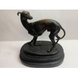 A bronze figure of a greyhound standing looking back over its shoulder, on black painted wood