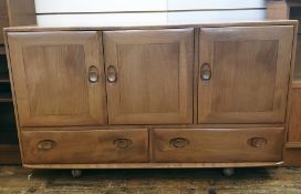 Light elm Ercol sideboard of three cupboard doors above two drawers, the whole raised on castors,