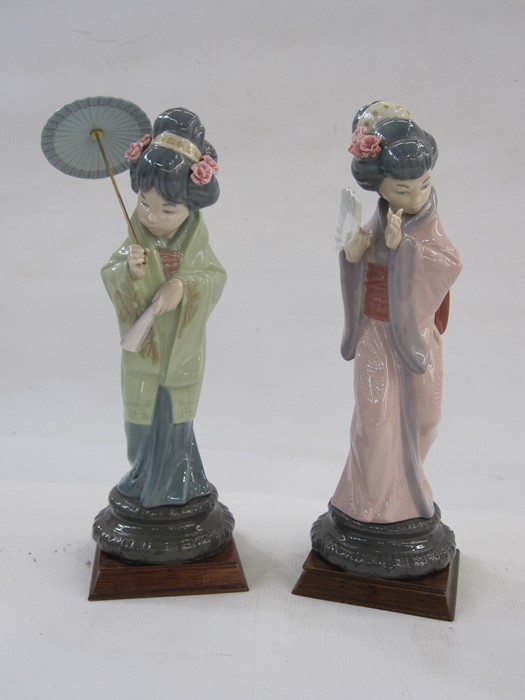 Two Lladro style Geisha figures holding umbrella and fan, one in green kimono, other in purple