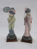 Two Lladro style Geisha figures holding umbrella and fan, one in green kimono, other in purple