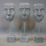 Three reconstituted stone masks on stands with block bases, H.35cm