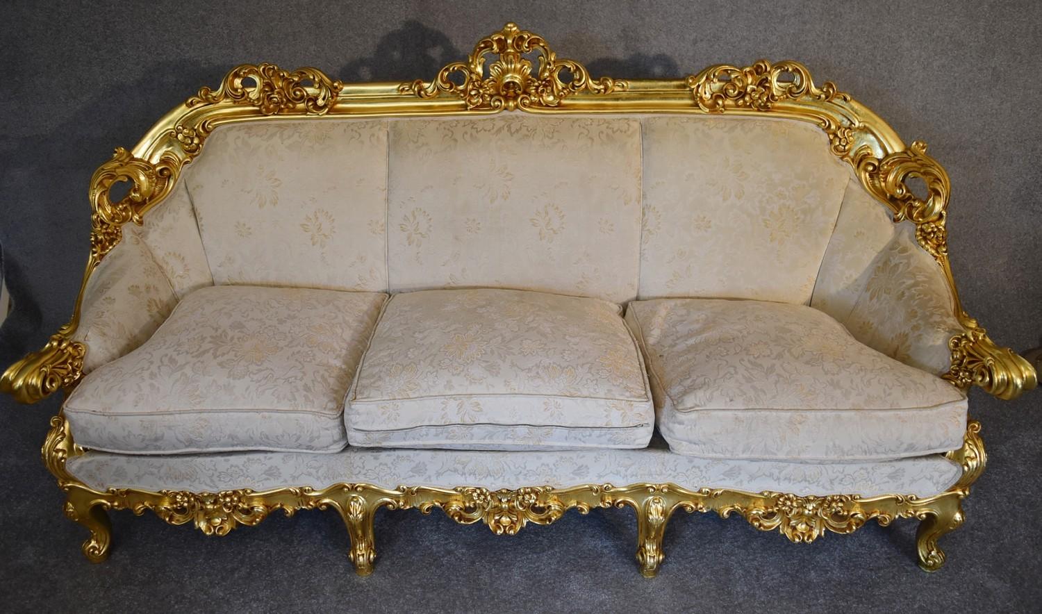 A heavily carved gilt framed Baroque style three cushion sofa with cream fabric and gold coloured