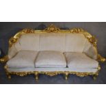 A heavily carved gilt framed Baroque style three cushion sofa with cream fabric and gold coloured