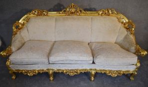 A heavily carved gilt framed Baroque style three cushion sofa with cream fabric and gold coloured