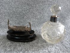 A silver collared cut glass perfume bottle and a silver wire boat on wooden stand. Perfume bottle