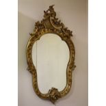 A DECORATIVE GILTWOOD CARVED WALL MIRROR, the shaped glass set within a carved Rococo style frame w
