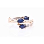 A yellow metal and triple sapphire crossover style ring, set with three mixed oval-cut sapphires and