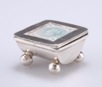 A SILVER METAL STAMP BOX, with hinged square lid having a glazed aperture and raised on four ball