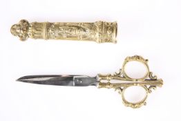 A PAIR OF VICTORIAN BRASS SEWING SCISSORS, in a scabbard cast with a Classical figure. 17.5cm