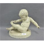 White painted plaster figure of a baby boy, modelled seated, pulling off his sock, on an oval plinth