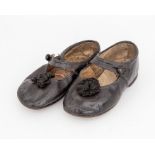 A pair of black leather children's shoes with a strap and button fastening, rosette detail on the