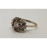 A precious white metal, diamond and emerald dress ring, set round brilliant cut diamond to centre