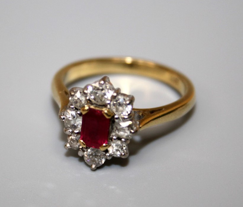 A ruby and diamond cluster ring, the step cut, cut cornered ruby in four claw mount above a border