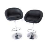 A pair of white metal and black leather tub bar stools, with foot rests, raised on a circular base.