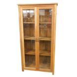 A light oak display cabinet, with a plain pediment over two bevelled glass inset doors, opening to