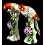 A pair of continental porcelain parrots, each standing on a simulated wooden perch decorated with pu
